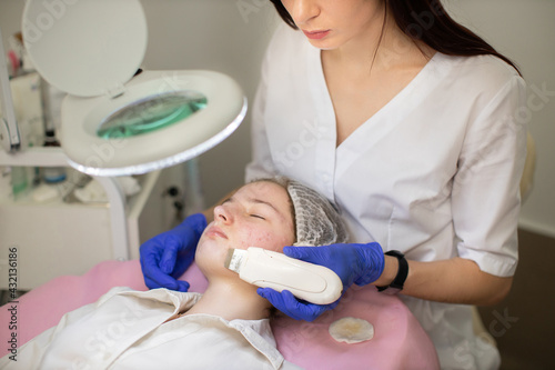Professional beautician using ultrasonic device in the hands and making procedure for cleansing the skin. Young teen girl with acne skin at modern cosmetology clinic