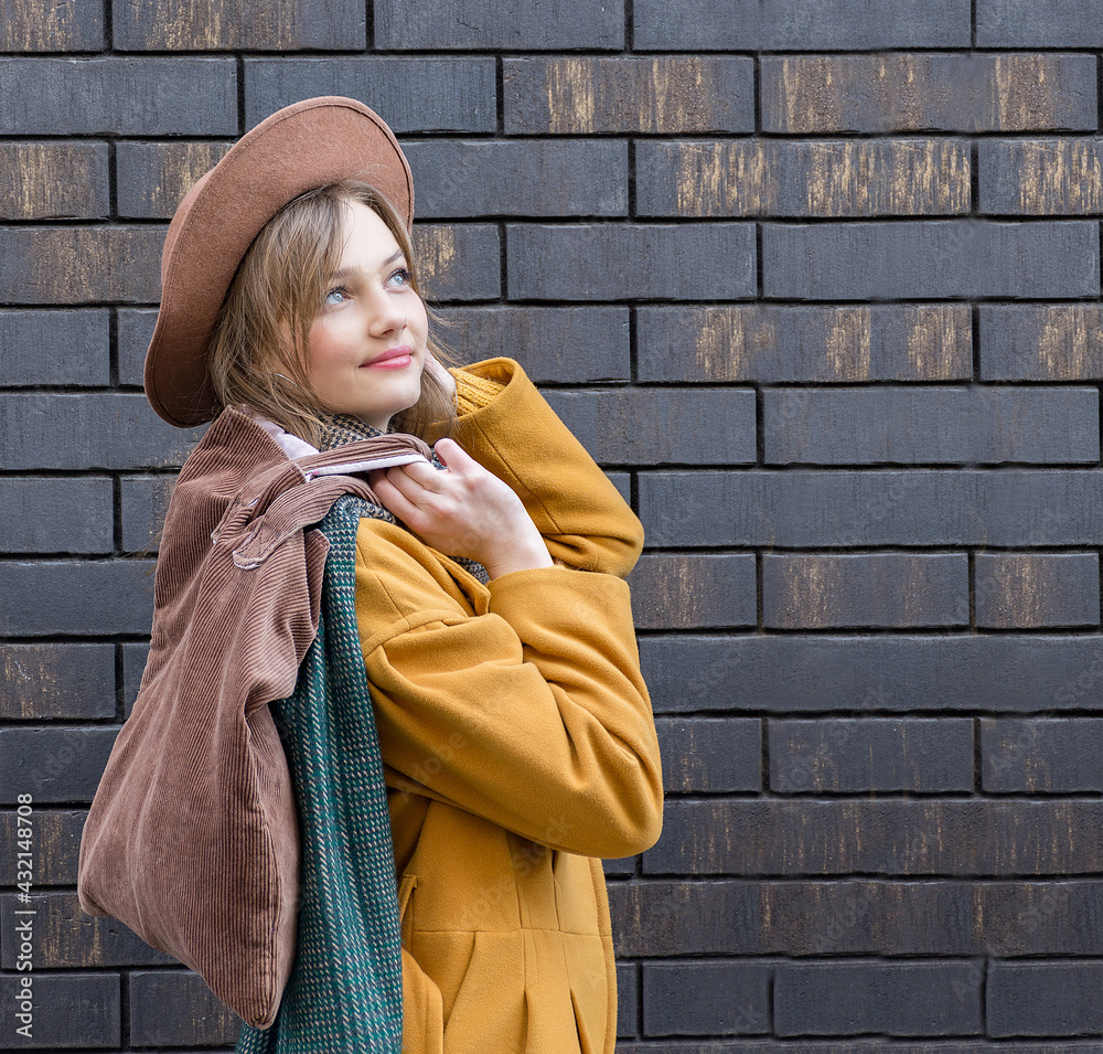 Naklejka premium Young woman holding a brown eco textile bag with space for mockup. Dressed in a yellow coat, hat, scarf. The concept of ecology or environmental protection. Ecological shopping. Style and fashion.