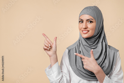 Young cheerful arabian muslim islamic woman in grey hijab pointing at copy space isolated on beige background. 30s Middle Eastern woman in hijab showing advertisement place photo