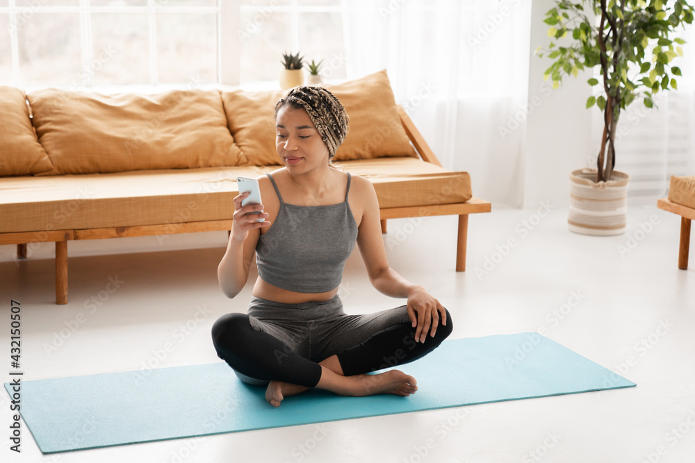 Fitness afro young girl in sporty clothes using smart phone during home sports training on mat indoors. Young african woman relaxing after home workout Fitness afro young girl in sporty clothes using smart phone during home sports training on mat indoors. Young african woman relaxing after home workout