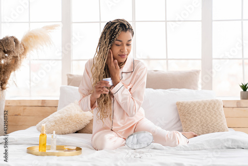 Beautiful young african woman in pyjamas applying moisturizing cream on her face sitting on the bed, taking care of her skin, body care, beautification concept photo