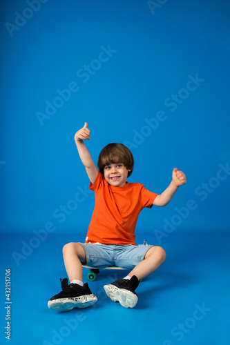 happy boy toddler sits on a skateboard and holds his hands up on a blue background with space for text