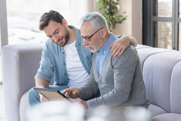 Caring loving adult son hugging embracing his old elderly senior father while he is showing his family photograph photo at home, telling stories of his youth. Happy father`s day! I love you, dad!