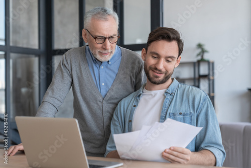 Caring loving adult son helping his old senior elderly father paying bills, counting money, doing paperwork at home together. Aid to elderly generation. Happy father`s day!