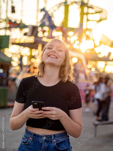 Mujer joven usando su móvil durante la puesta de sol en una feria o parque de atracciones