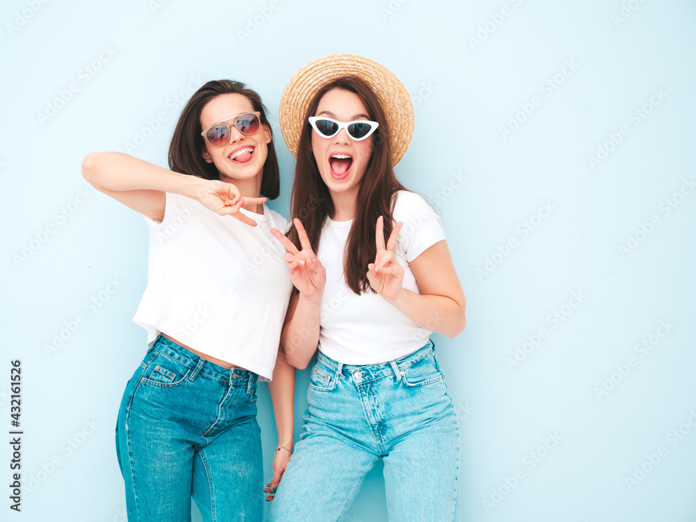 Two young beautiful smiling hipster female in trendy summer white t-shirt and jeans clothes.Sexy carefree women posing near light blue wall in studio.Positive models in hats.Showing peace sign