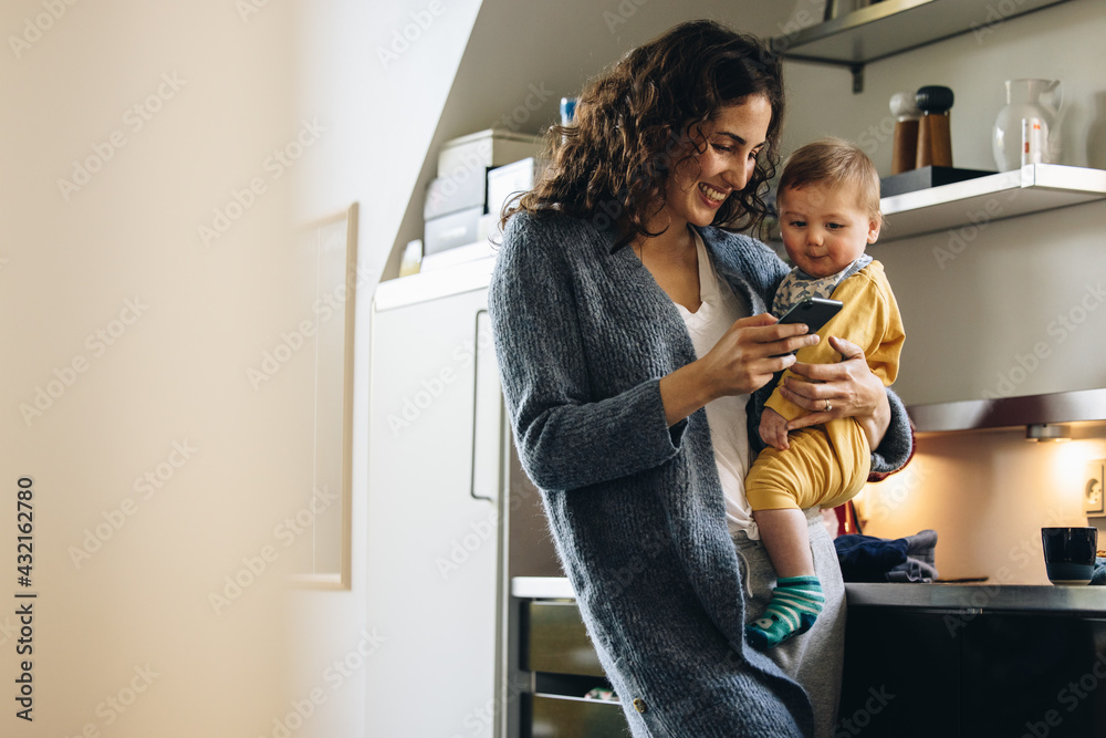 © Jacob Lund - Smiling woman with baby texting on phone at home