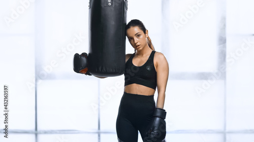 exhausted sportswoman in boxing gloves and sportswear leaning on punching bag in gym. © LIGHTFIELD STUDIOS
