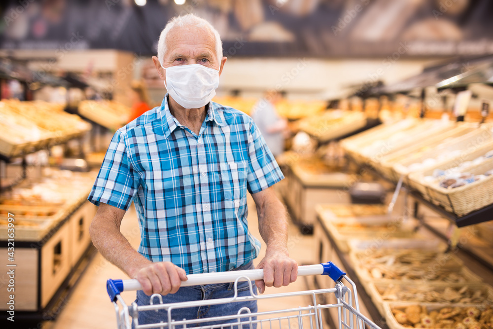 older european man wearing mask and gloves with covid protection chooses buns and bread in supermarket bakery