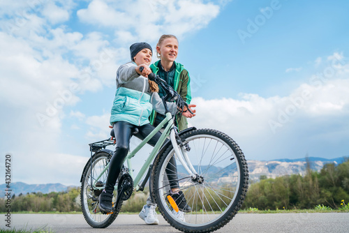 Brother helping to sister, supporting her and teaching doing first steps in riding. Kids on a bicycle path with wide blue cloudy sky background. Happy childhood concept image.