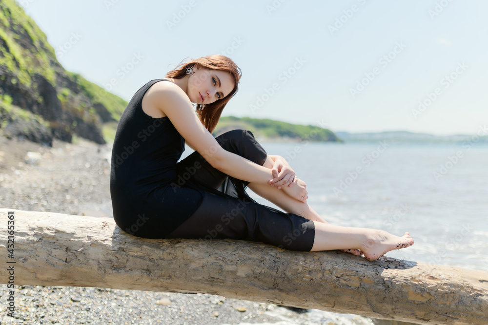 Young woman in black dress sitting on sand and enjoys sea.