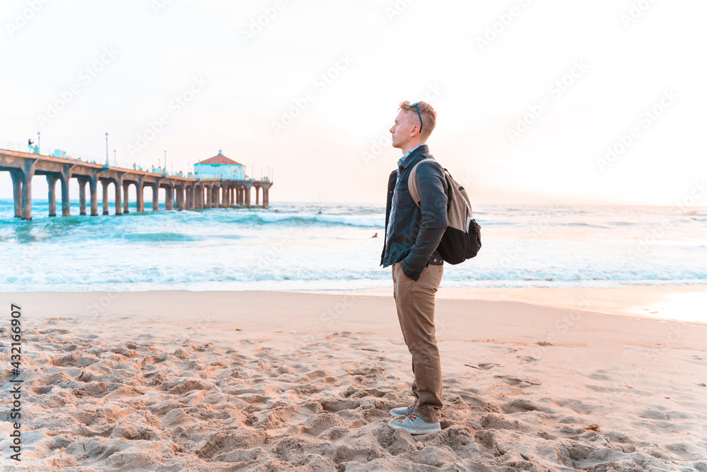 A young man with a backpack walks on the beach in Los Angeles with a bright sunset A young man with a backpack walks on the beach in Los Angeles with a bright sunset