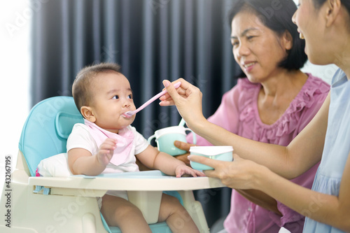 Young asian mother and grandmother feeding her baby with fruit puree.  Her son sitting on highchair in living room.