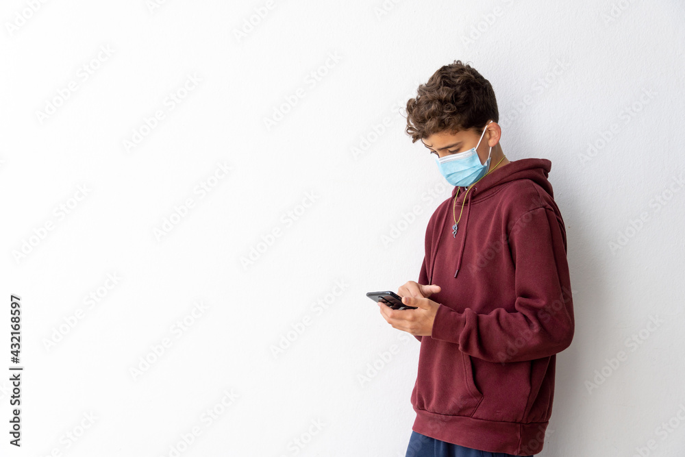 boy with mask using smartphone leaning against a wall with white background