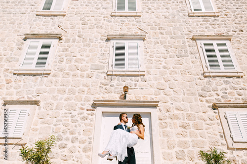 Groom holding bride in his arms against the stone wall of the building on a bright sunny day. © Nadtochiy
