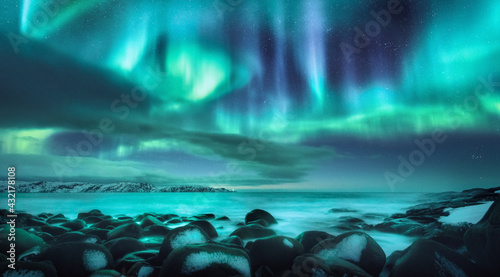 Northern lights. Aurora borealis over ocean in Teriberka, Russia. Starry sky with polar lights and clouds. Night winter landscape with bright aurora, stars, sea, snowy stones in blurred water. Travel © den-belitsky
