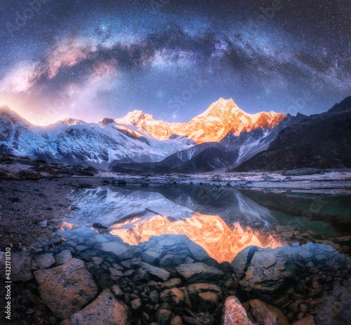 Milky Way over snowy mountains and lake at night. Landscape with snow covered high rocks, starry sky, stones, reflection in clear water in Nepal. Sky with stars. Bright milky way in Himalayas. Space 