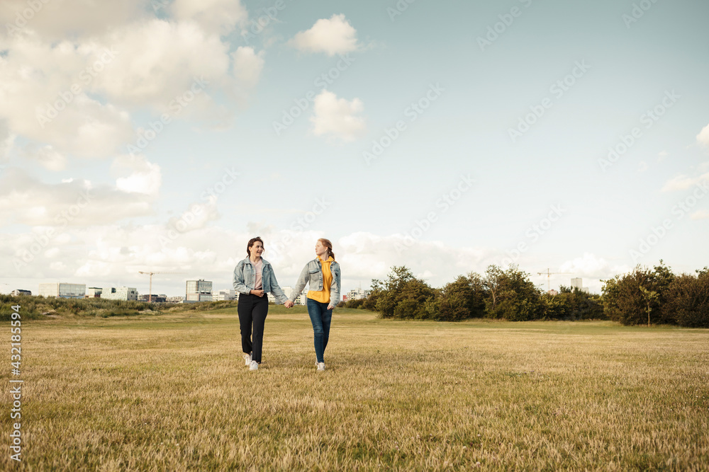 Female friends holding hands while walking in park against sky Female friends holding hands while walking in park against sky