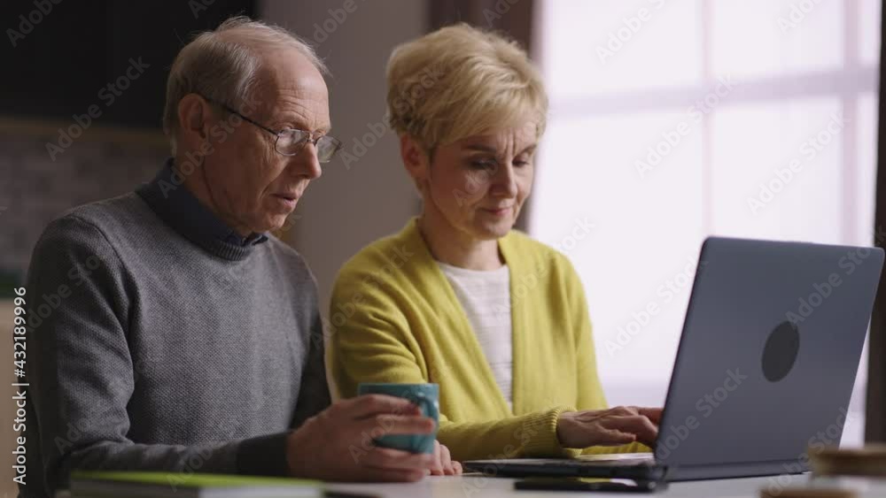 elderly spouses are using laptop together, sitting at home kitchen at weekend, shopping online or communicating in social nets elderly spouses are using laptop together, sitting at home kitchen at weekend, shopping online or communicating in social nets