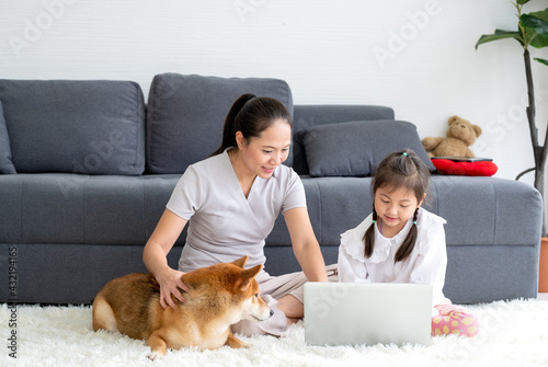 Shiba inu dog interest in little girl has fun with laptop and also sit near her mother at living room during stay at home to prevent infection of disease pandemic outside. © narong