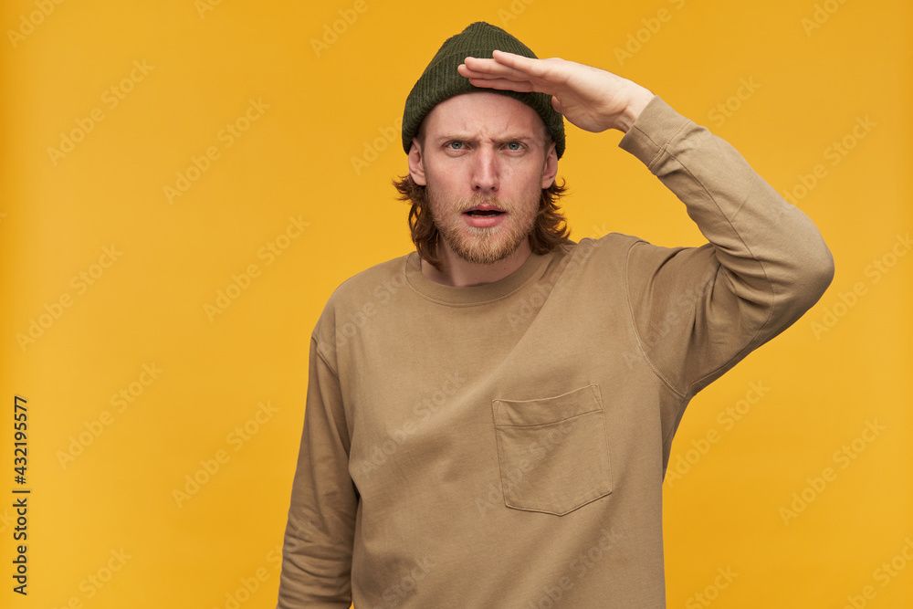 Portrait of serious male with blond hair and beard. Wearing green beanie and beige sweater. Holds palm next to forehead and gaze into distance. Watching at the camera isolated over yellow background