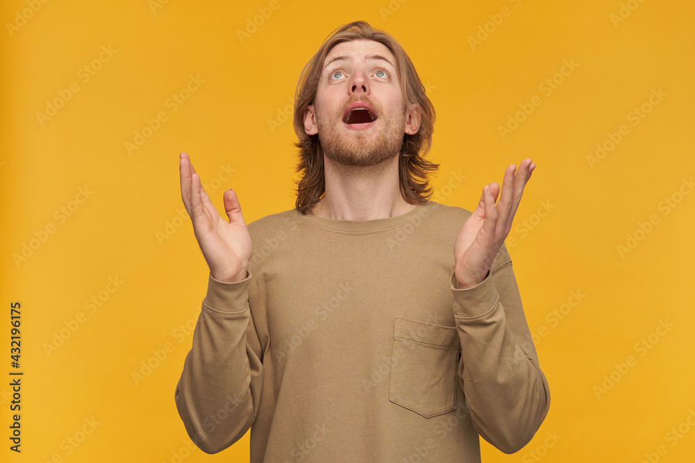 Young blessed man, positive guy with blond hair, beard and mustache. Wearing beige sweater. Raising his palms and watching admiringly up at copy space, isolated over yellow background Young blessed man, positive guy with blond hair, beard and mustache. Wearing beige sweater. Raising his palms and watching admiringly up at copy space, isolated over yellow background