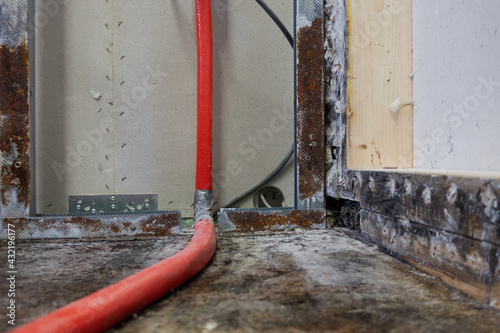 Drywall (Trockenbau) with rusty metal rails and red isolated water pipe. Mold and water stains on brown wood floor. Insurance claim and construction drying.