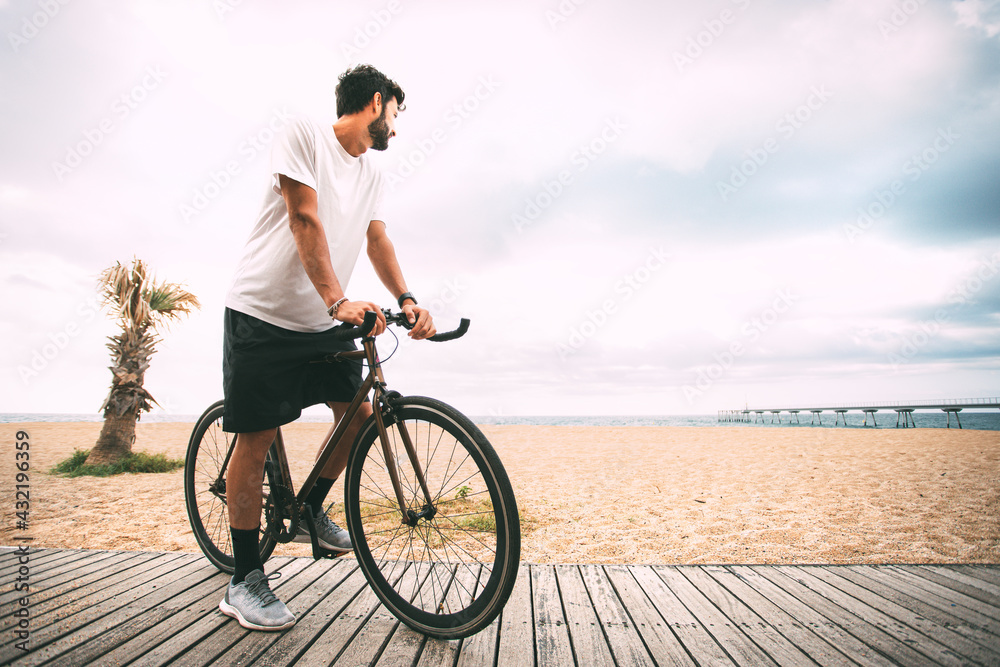 Fototapeta premium boy on a bike on a wooden walkway on the beach looking at the sea and a bridge in the background. Clear sky background with copy space. sport concept. urban cycling concept. technology concept.