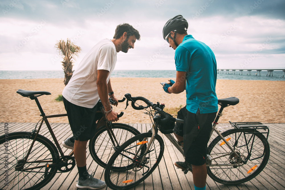 Obraz premium Cyclists on a bike on a wooden walkway on the beach with the sea and a bridge in the background looking at the mobile phone. Clear sky background with copy space. sport concept. urban cycling concept
