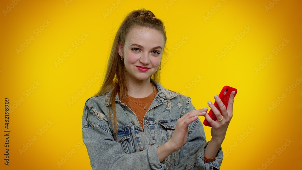 Teenager student girl in denim jacket using mobile phone typing new post on web, sms message, browsing, addiction of social networks. Young fashionable woman indoors studio shot on yellow background Teenager student girl in denim jacket using mobile phone typing new post on web, sms message, browsing, addiction of social networks. Young fashionable woman indoors studio shot on yellow background