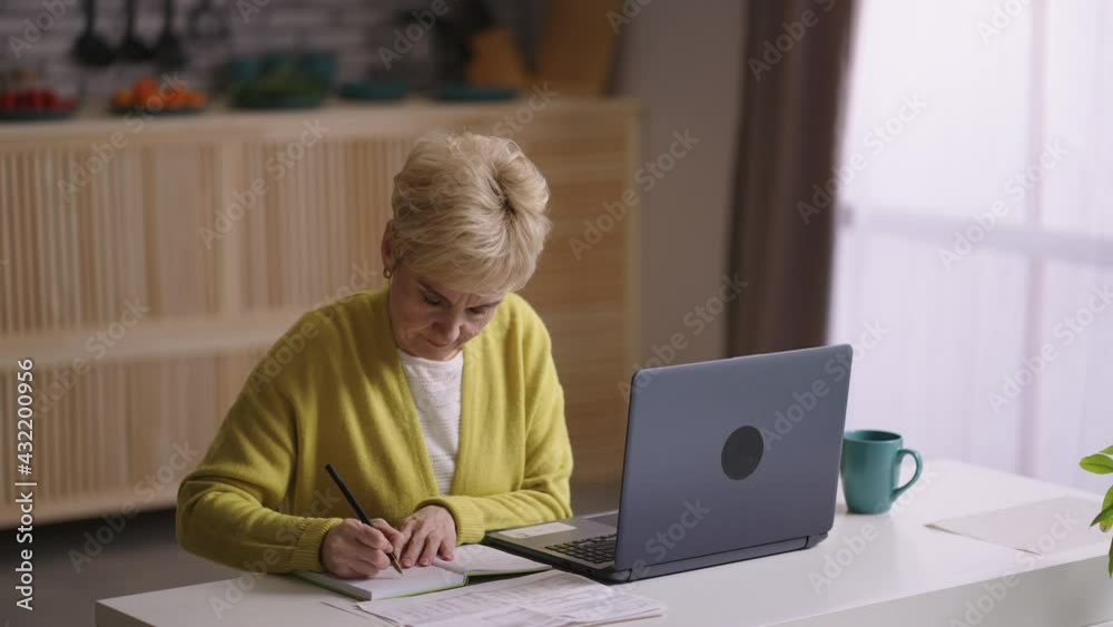 old woman is learning online, sitting at home kitchen with laptop, looking for information in internet and making notes in notebook old woman is learning online, sitting at home kitchen with laptop, looking for information in internet and making notes in notebook