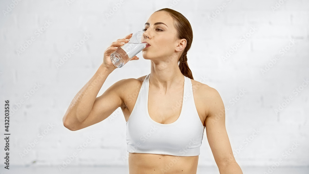 Sportswoman in white sportswear drinking water near white wall. - obrazy, fototapety, plakaty 