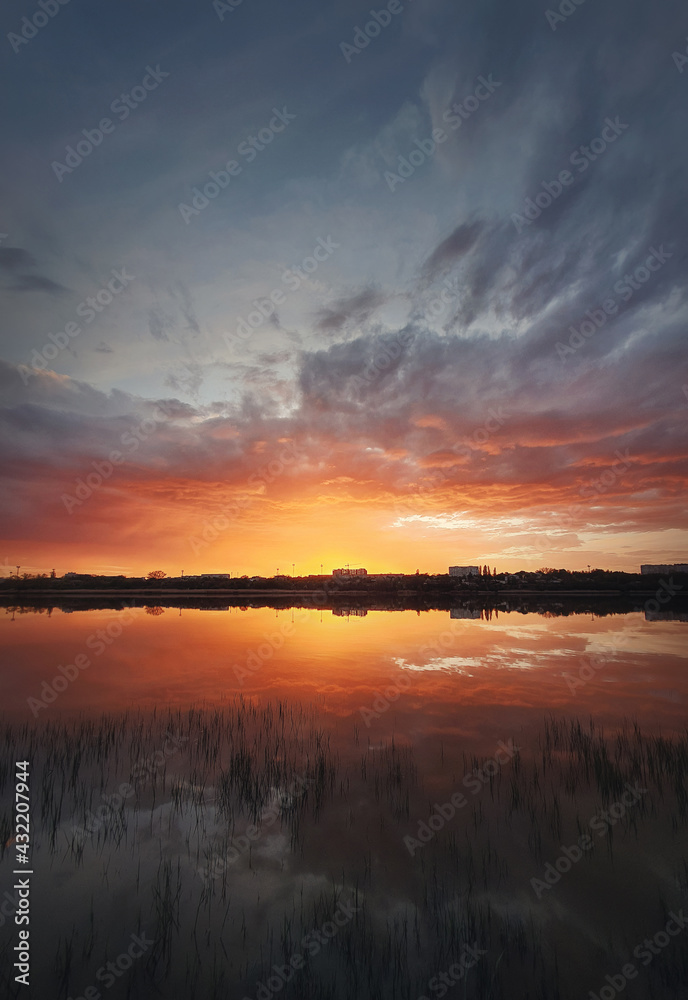 Fototapeta premium Fascinating sunset reflecting on the lake surface. Idyllic landscape, vertical background. Silent and tranquil evening scene with colorful sky, reed vegetation in the pond and a city at the horizon