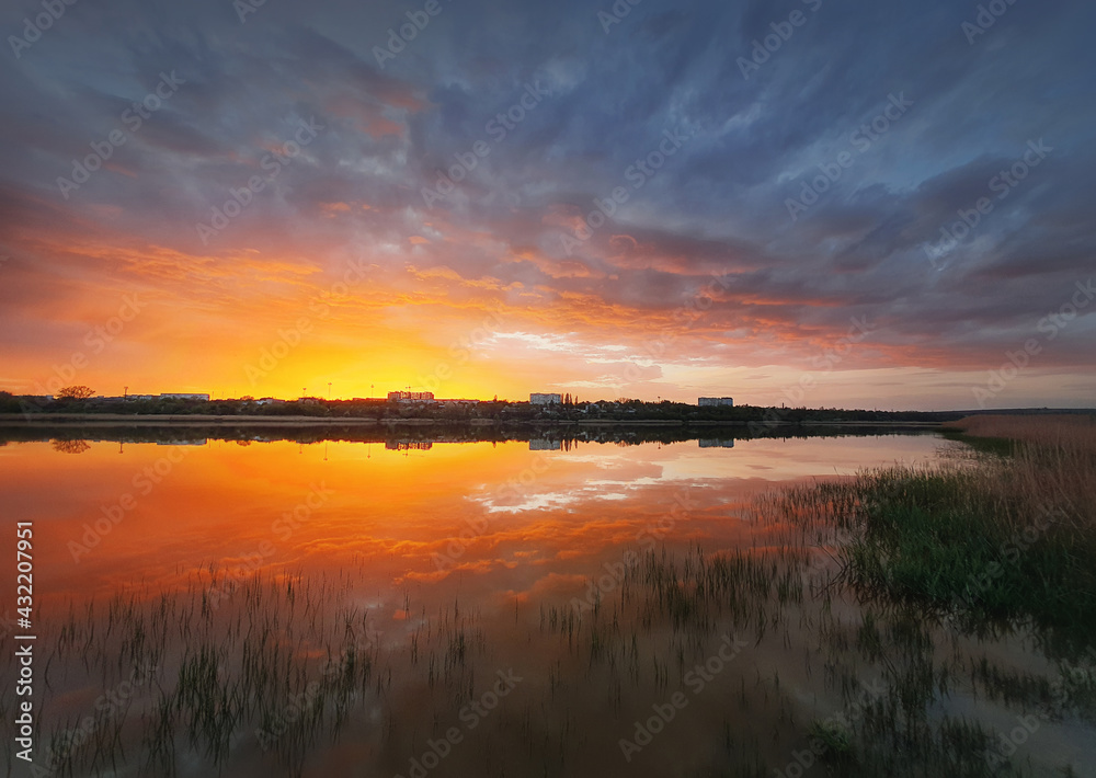 Naklejka premium Fascinating sunset reflecting on the lake surface. Idyllic landscape, vertical background. Silent and tranquil evening scene with colorful sky, reed vegetation in the pond and a city at the horizon