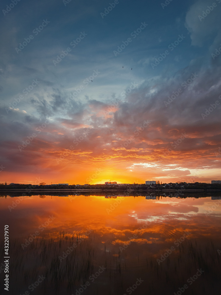 Fascinating sunset reflecting on the lake surface. Idyllic landscape, vertical background. Silent and tranquil evening scene with colorful sky, reed vegetation in the pond and a city at the horizon