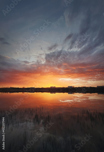 Fascinating sunset reflecting on the lake surface. Idyllic landscape, vertical background. Silent and tranquil evening scene with colorful sky, reed vegetation in the pond and a city at the horizon © psychoshadow