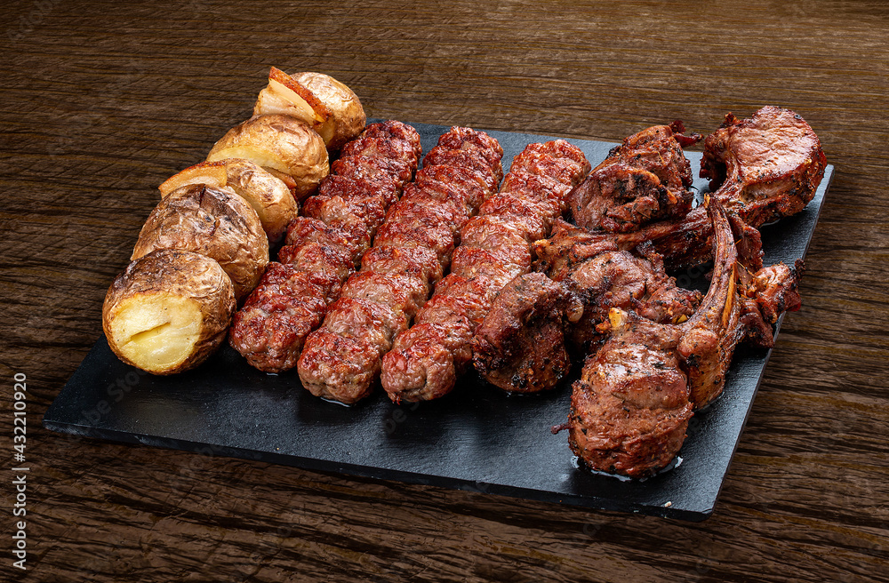 A selection of various barbecued gourmet meats on a black board with a rustic timber background. Rack of lamb, potatoes, kebab