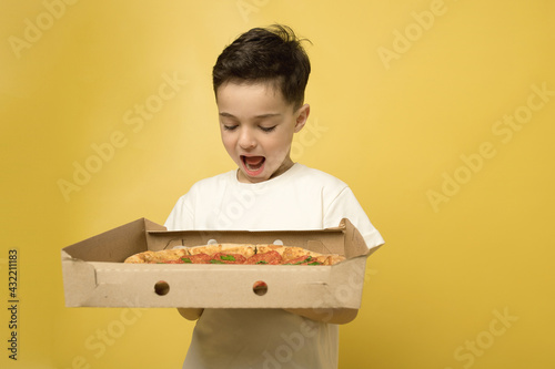 Brunet child posing with pizza in delivery box isolated on yellow background. Funny hungry boy is holding big pizza in a to go box. Caucasian. Fast food, junk food, children love pizza concept. 