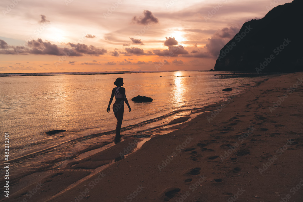 Slender woman walking along ocean shore at amazing orange sunset