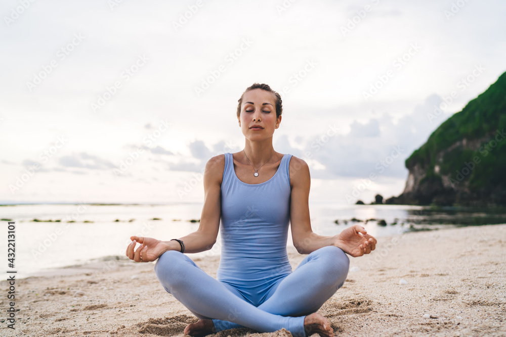 Caucasian female with closed eyes practicing yoga at coastline reaching healthy lifestyle and wellness, calm woman with flexible body sitting in lotus pose enjoying asana time for mindfulness