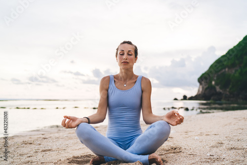 Caucasian female with closed eyes practicing yoga at coastline reaching healthy lifestyle and wellness, calm woman with flexible body sitting in lotus pose enjoying asana time for mindfulness