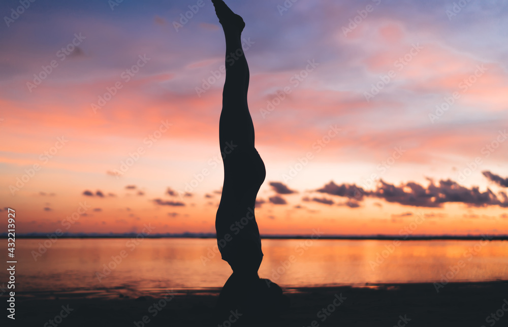 Side silhouette view of slim female athlete doing headstand exercise during evening workout near ocean and colorful horizon, fit girl practice strength and body balance keeping healthy lifestyle
