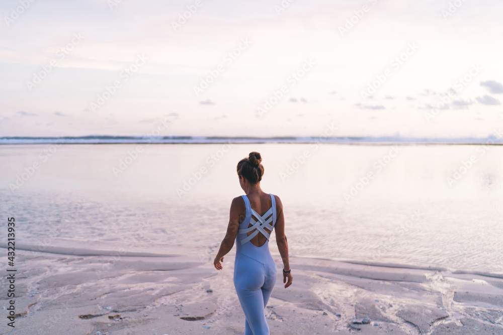 Back view of slim muscular fit girl dressed in sportive tracksuit walking at seashore coastline during morning time ready for active workout,female athlete spending time at calm environment with ocean