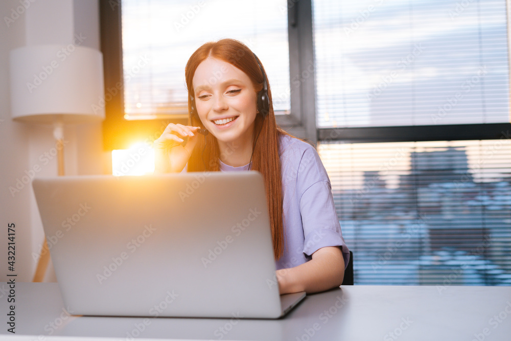 Close-up face of friendly young woman operator using headset and laptop during customer support at home office. Young redhead female student communicating online by video call on background of window. Close-up face of friendly young woman operator using headset and laptop during customer support at home office. Young redhead female student communicating online by video call on background of window.