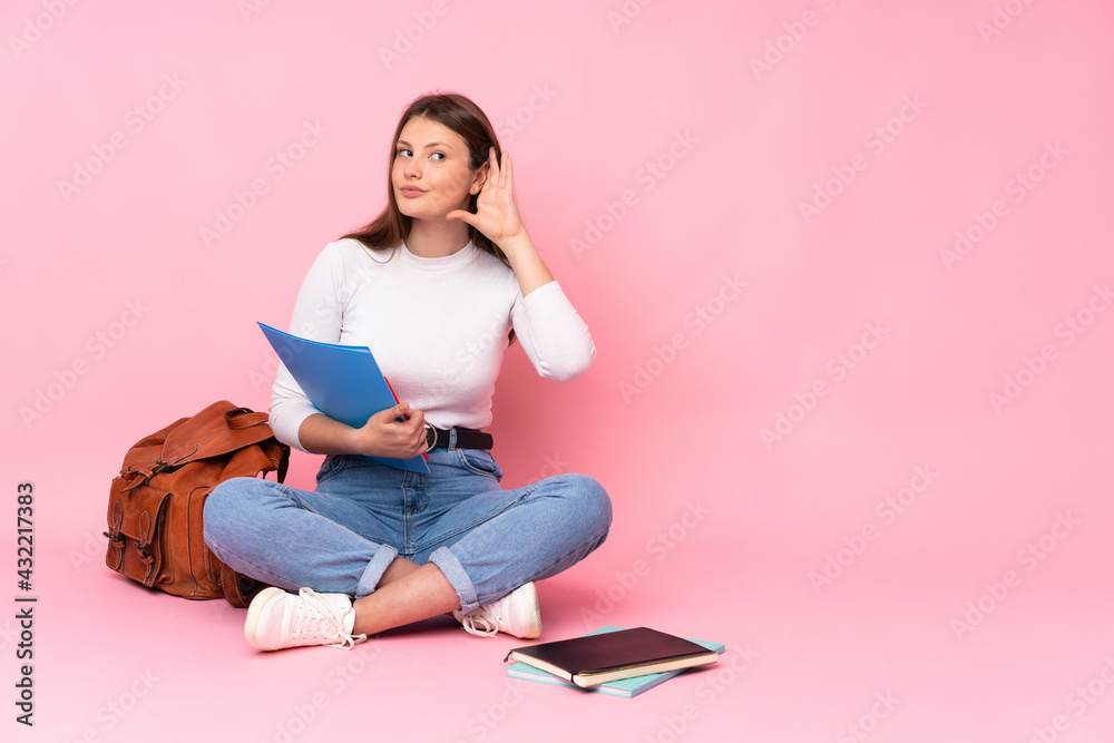 Teenager caucasian student girl sitting on the floor isolated on pink background listening to something by putting hand on the ear Teenager caucasian student girl sitting on the floor isolated on pink background listening to something by putting hand on the ear