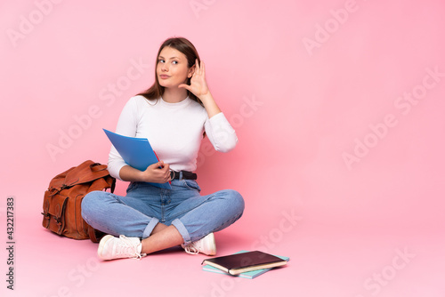 Teenager caucasian student girl sitting on the floor isolated on pink background listening to something by putting hand on the ear