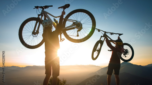 Two cheerful woman lifts his bicycle above his head at sunset after a winning mountain biking ride.