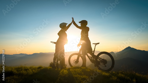 Two happy woman high five over the sunset after a successful mountain biking trip in the mountains. Celebrate a cross country cycling journey.