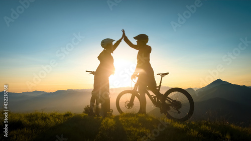 Two happy woman high five over the sunset after a successful mountain biking trip in the mountains. Celebrate a cross country cycling journey. photo