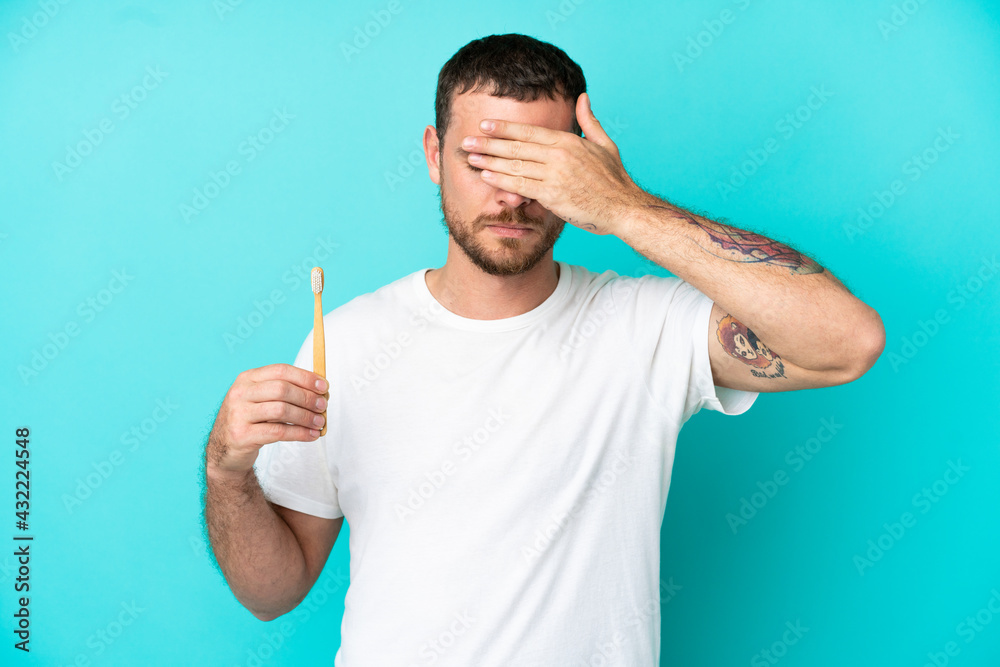 Young Brazilian man brushing teeth isolated on blue background covering eyes by hands. Do not want to see something